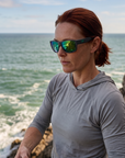 Woman, 35, wearing Peppers Big Kahuna sunglasses in Dark Grey Frame with Green Diamond Mirror Lens, on a sea cliff above the water