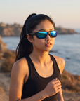 Woman, 21, wearing Peppers Cutthroat sunglasses in Matte Black Frame with Brown Ice Blue Mirror Lens, on a coastal path above the ocean