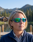 Woman, 43, wearing Peppers Cutthroat sunglasses in Matte Grey Frame with Emerald Green Mirror Lens, at a mountain-lake lodge dock