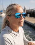 Woman, 45, wearing Peppers Defender sunglasses in Matte Grey Frame with Blue Revo Lens, on a charter boat deck in a fishing harbour