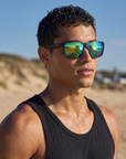 Man, 24, wearing Peppers Low Rider sunglasses in Matte Black Frame with Green Revo Lens, at a surf beach