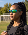 Woman, 37, wearing Peppers Ricochet sunglasses in Shiny Green / Silver Frame with Green Diamond Mirror Lens, beside an empty park sports field