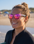 Woman, 23, wearing Peppers Sailfish sunglasses in Shiny Pink with Blue Pattern Temples with Pink Mirror Lens, on a surf beach