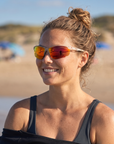 Woman, 23, wearing Peppers Three Wood sunglasses in Matte White Frame with Red Revo PC Lens, on a surf beach