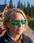 Woman, 43, wearing Peppers Anchor sunglasses in Shiny Crystal Grey Frame with Smoke Emerald Green Mirror Lens, at a mountain-lake lodge dock