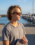Man, 22, wearing Peppers Beachcomber sunglasses in Matte Black Frame with Brown Fire Red Mirror Lens, in a harbour car park