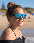 Woman, 23, wearing Peppers Big Kahuna sunglasses in Matte Black Frame with Blue Mirror Lens, on a surf beach
