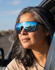 Woman, 48, wearing Peppers Cool Driver sunglasses in Matte Black Frame with Blue Revo Mirror, on a seaside road