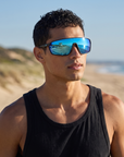Man, 24, wearing Peppers Cool Driver sunglasses in Matte Black Frame with Blue Revo Mirror, at a surf beach