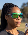 Woman, 37, wearing Peppers Cool Driver sunglasses in Matte Grey Frame with Green Revo Lens, beside an empty park sports field