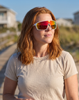 Woman, 31, wearing Peppers Cool Driver sunglasses in Matte White Frame with Red Revo PC Lens, on a beach boardwalk