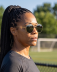 Woman, 37, wearing Peppers Django sunglasses in Gold Frame with G-15 Lens, beside an empty park sports field