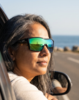 Woman, 48, wearing Peppers Enzo sunglasses in Smoke Crystal Frame with Green Diamond Mirror Lens, on a seaside road