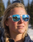 Woman, 27, wearing Peppers Grand Teton sunglasses in Matte Crystal Frame with Blue Revo Lens, at the base of a ski area