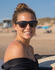 Woman, 23, wearing Peppers Grand Teton sunglasses in Matte Crystal Clear to Black Frame with Smoke Lens, on a surf beach