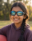 Woman, 10, wearing Peppers Kickball sunglasses in Black / Snow Camo with Blue Mirror Lens, at an empty park playing field