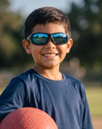 Man, 11, wearing Peppers Kickball sunglasses in Black / Snow Camo with Blue Mirror Lens, at an empty park playing field
