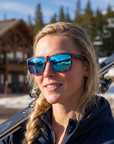 Woman, 27, wearing Peppers Low Rider sunglasses in Matte Tort Frame with Blue Revo Lens, at the base of a ski area