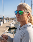 Woman, 45, wearing Peppers Mountain Madness sunglasses in Shiny Crystal Grey Frame with Green Diamond Mirror Lens, on a charter boat deck in a fishing harbour