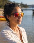 Woman, 29, wearing Peppers Mountain Madness sunglasses in Matte Crystal Clear to Black Fade Frame with Red Diamond Mirror Lens, at a calm bay