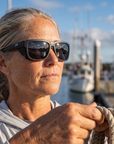 Woman, 45, wearing Peppers Sea Dweller 2.0 sunglasses in Matte Black Frame with Smoke Lens, on a charter boat deck in a fishing harbour