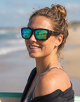 Woman, 23, wearing Peppers Sea Dweller 2.0 sunglasses in Matte Dark Green / Silver Frame with Green Diamond Mirror Lens, on a surf beach
