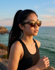 Woman, 21, wearing Peppers Seawitch sunglasses in Matte Black To Tortoise Frame with Brown Lens, on a coastal path above the ocean
