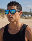 Man, 24, wearing Peppers Whaler sunglasses in Matte Black Frame with Blue Diamond Mirror Lens, at a surf beach