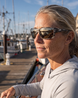 Woman, 45, wearing Peppers Whaler sunglasses in Grey Striped Frame with Brown Silver Flash Lens, on a charter boat deck in a fishing harbour